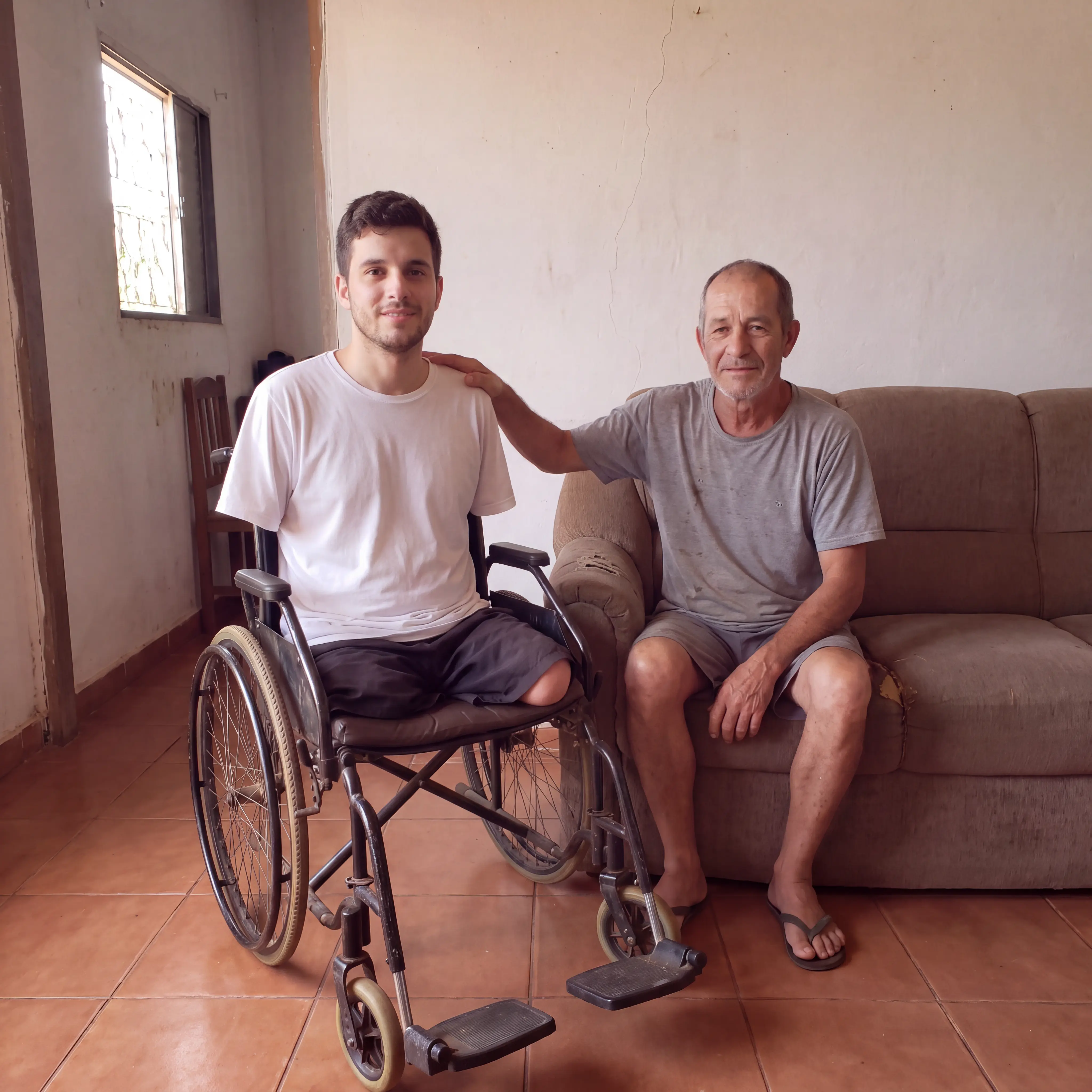 Bruno em sua cadeira de rodas ao lado do pai, sentados dentro de casa
